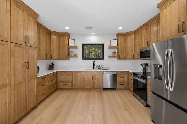 The Salem House kitchen with natural wood cabinets, white countertops, stainless steel appliances, and a window overlooking the woods.